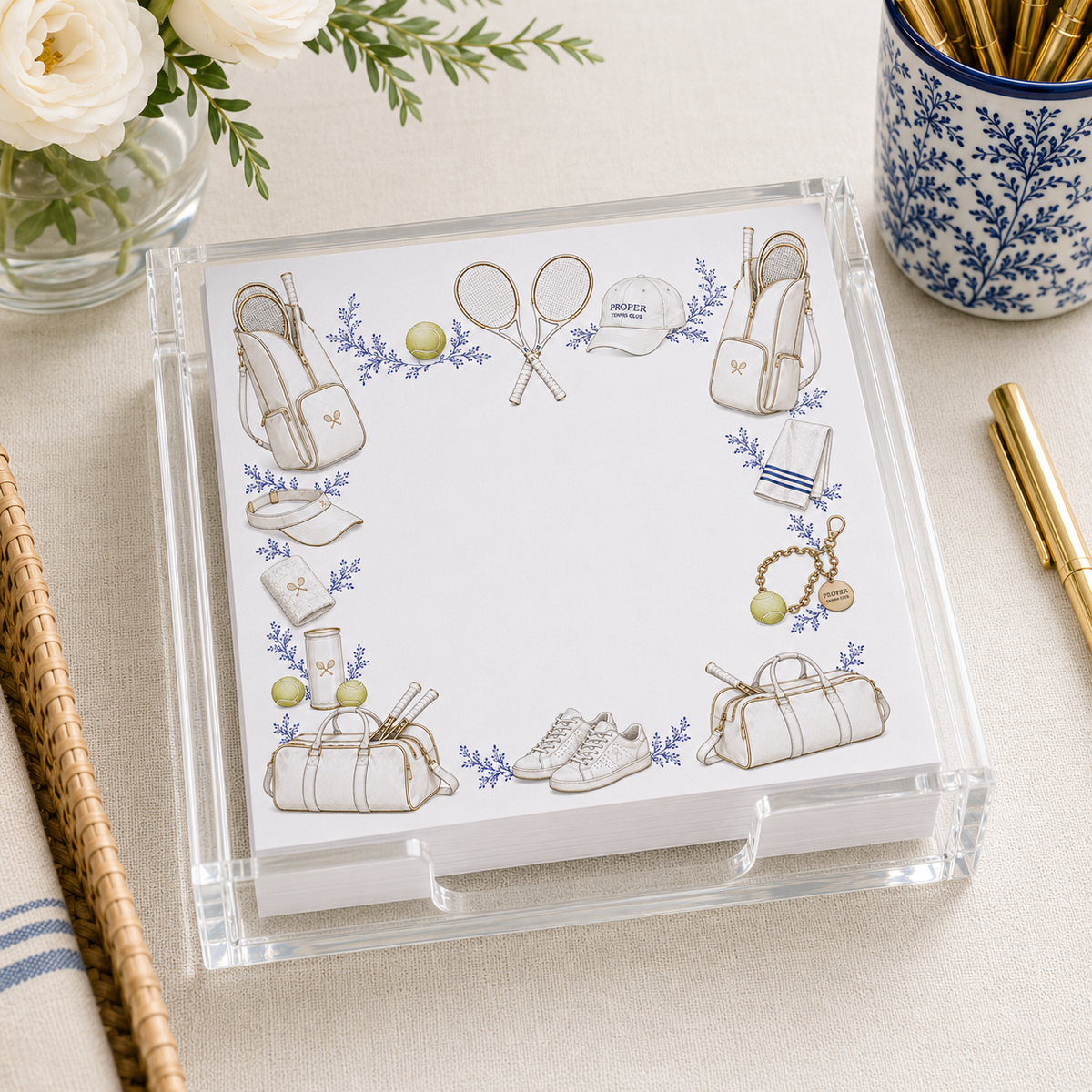 Clear acrylic tray holding tennis-illustrated notepads in a styled desk scene with gold pen and white roses